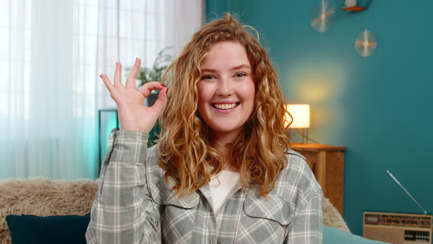 Young woman at home showing OK hand sign with confident look expressing agreement and positive attitude. Caucasian redhead girl on sofa smiling proudly, emotional strength and self-assured posture