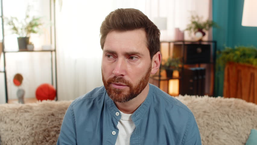Young man at home sits on sofa, squinting looking at camera with suspicion, doubting what he sees. Caucasian guy looks skeptical narrowing eyes in disbelief feeling unsure about unexpected information
