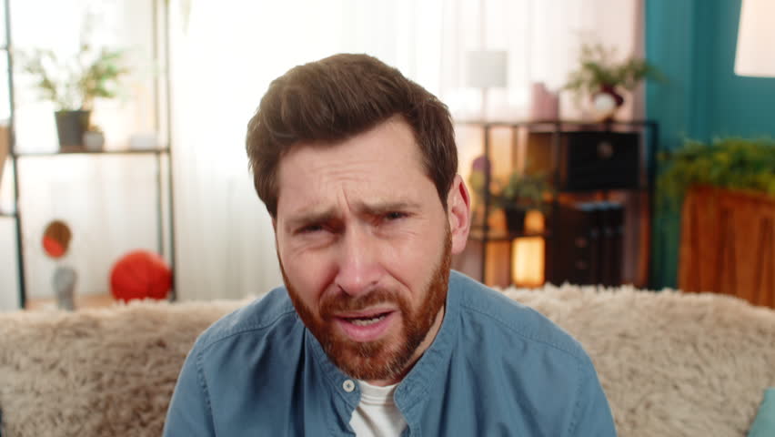 Young man at home sits on sofa, squinting looking at camera with suspicion, doubting what he sees. Caucasian guy looks skeptical narrowing eyes in disbelief feeling unsure about unexpected information