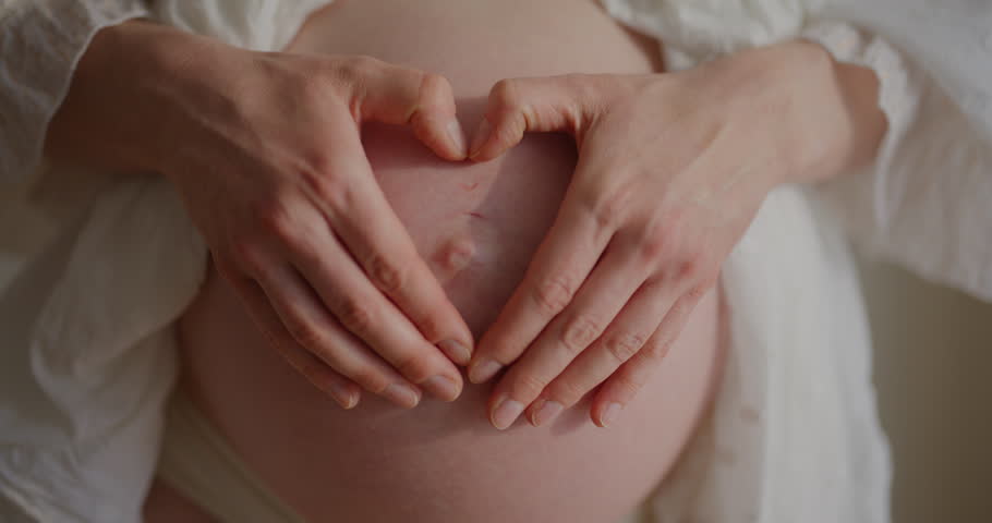 Close-up of a pregnant woman's belly in the 9th month, with her hands forming a heart shape.