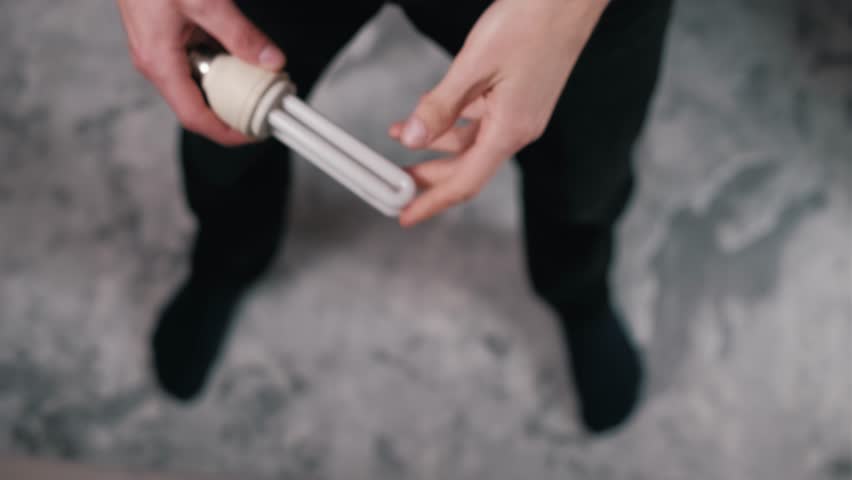 Overhead perspective captures two male hands carefully examining a compact fluorescent light bulb. Detailed view shows inspection of the energy-efficient CFL lamp against a blurred floor.