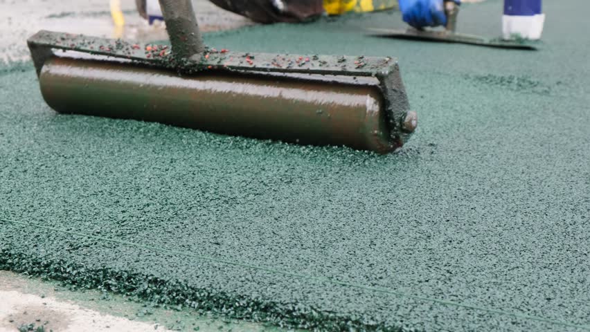 Construction workers manually install EPDM granules on an outdoor playground. One builder uses a metal roller to press the rubber layer. The soft surface reduces injury risk for children.