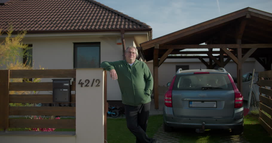 Man leaning on a fence beside a residential mailbox with a suburban house and parked car in the background, exuding a calm and approachable presence
