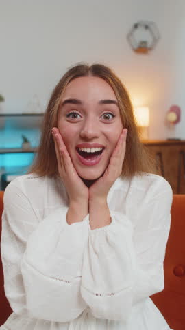 Oh my God, Wow. Young woman surprised looking at camera with big eyes, shocked by sudden victory, good win news, celebrating at home. Excited amazed girl indoors in living room on couch. Vertical