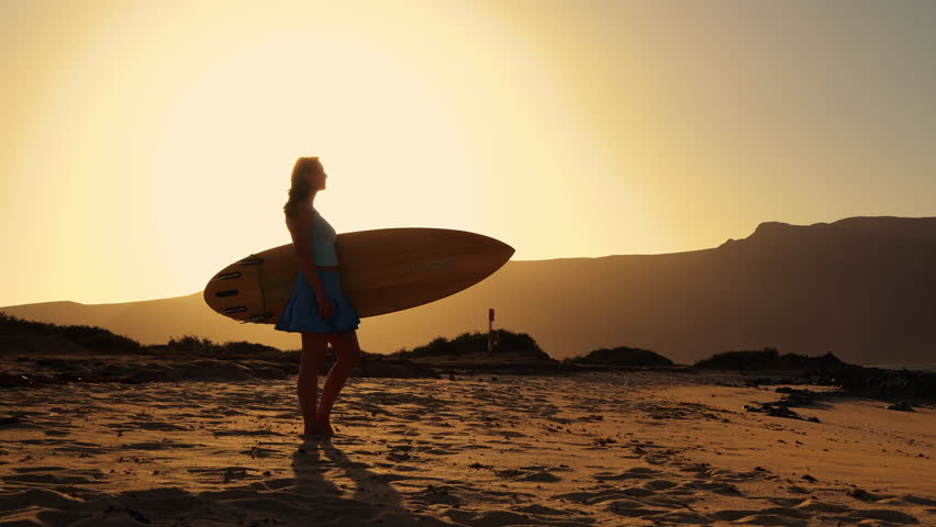 LENS FLARE, SILHOUETTE: Young woman stands on beach at sunset holding a wooden surfboard. She is checking the waves at a surf spot before heading into water for an evening session on Canary Islands.