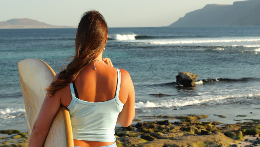 SLOW MOTION, CLOSE UP: Back view of a female surfer staring at beautiful breaking waves. Offshore wind grooms the waves and ruffles long hair of a young woman heading for a surf session on Lanzarote.