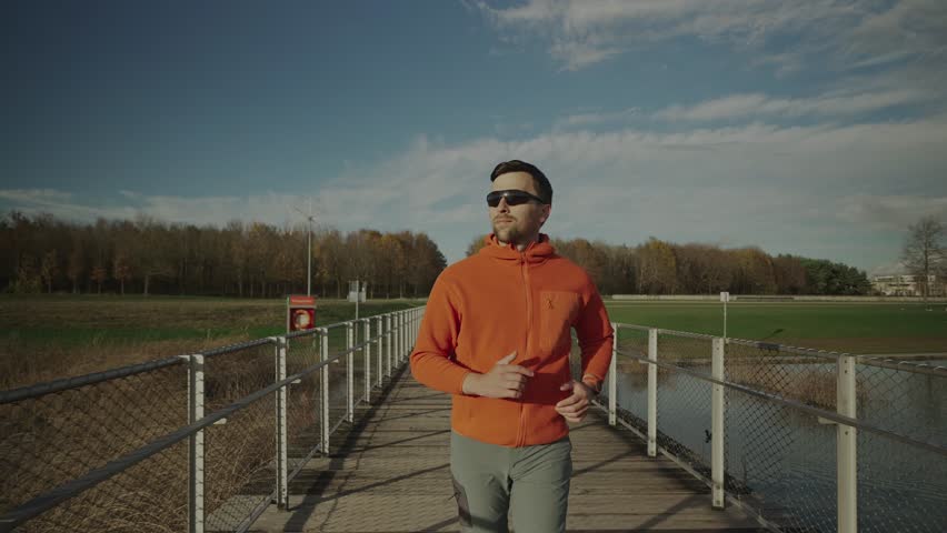 Cross country jogging in countryside. Man in orange sweatshirt running across wooden bridge in national park. Runner running over a wildlife trail wooden footbridge. Trail run in cold fall weather.  - Powered by Shutterstock - Get 15% off with code: PIKWIZARD15