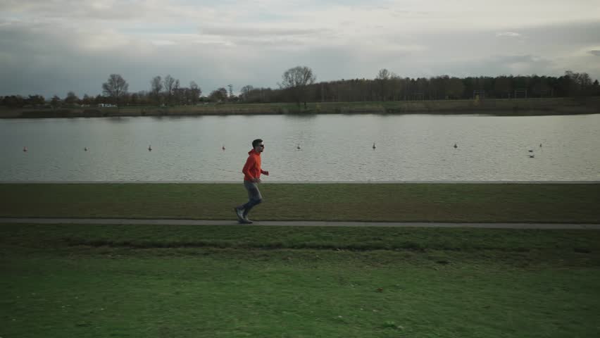 Sporty man jogging along footpath along lake in park in Germany, Munich, Germany, in autumn. Male running beside a pond in the fall. Outdoor exercise by the water. People and sport concept. 