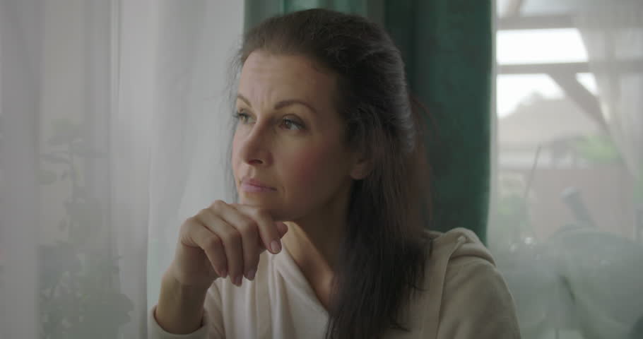 Woman sitting on a cozy sofa near a window, resting her arm on a cushion, looking outside with a thoughtful expression in a serene living room setting