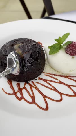 Close-up of a spoon breaking into a molten chocolate cake, revealing rich flowing lava. Served with vanilla ice cream, raspberry and mint on a white plate. Elegant gourmet dessert.