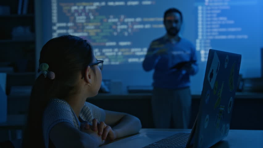 Medium closeup of Chinese schoolgirl sitting at desk at coding lesson, listening to teacher explaining new algorithm at whiteboard in blue projector light, then typing on laptop while performing task