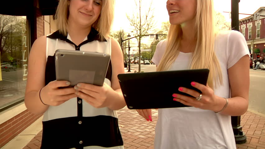 Two young women enjoy using their tablet PCs while walking in a town.