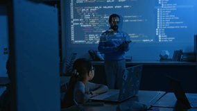 Full shot of male programming teacher demonstrating computer code on blue projector screen in dark classroom to school students with laptops, pointing at commands, girl raising hand to ask question - Powered by Shutterstock - Get 15% off with code: PIKWIZARD15