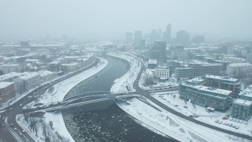 Drone Shot of River Neris winds gracefully through Vilnius, its icy banks blanketed in snow as a gentle blizzard falls from the grey sky