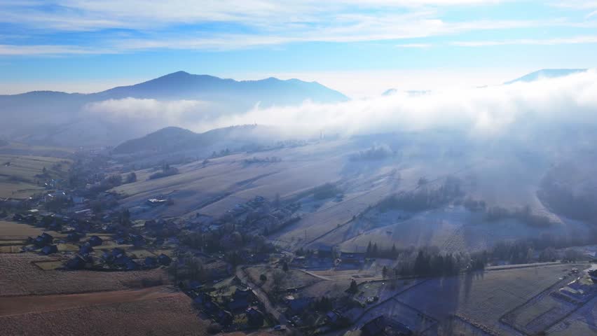 Aerial hyperlapse video of rolling hills, fog drifting over frosty fields, and scattered rural villages under a bright blue sky.