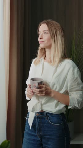 Young thoughtful beautiful woman drinking tea and looking through window while standing on windowsill at home. Footage of attractive girl tasting drink on background of modern living room. Indoors