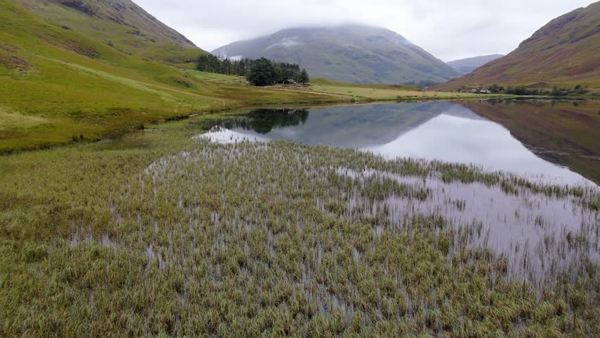 Aerial drone footage of Loch Achtriochtan in the Glencoe mountain range in the Scottish Highlands, taken in summer on a clear but misty day, morning time.