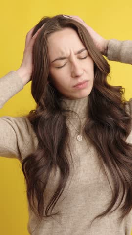 Woman holding her head, showing signs of discomfort and pain, indicating a severe headache or stress against a vibrant yellow backdrop