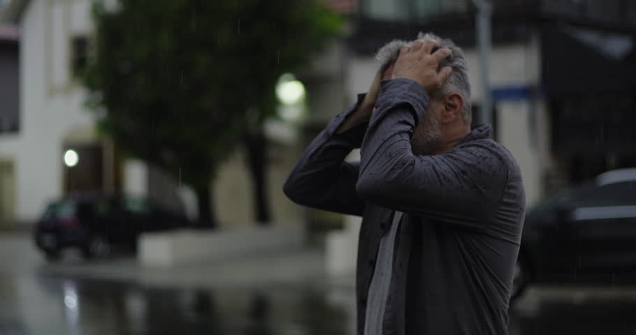 Middle aged man outdoors in rain with hand on head, eyes shut in visible frustration or mental exhaustion