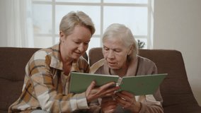 Elderly mother and middle-aged daughter spending their free time together. Two happy women looking at photos in album while sitting on sofa in room and experiencing positive emotions about memory. - Powered by Shutterstock - Get 15% off with code: PIKWIZARD15