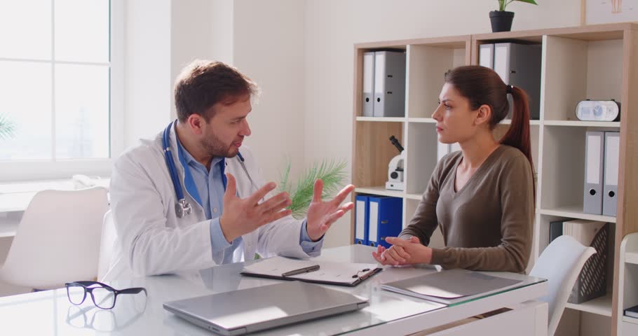 Male doctor in medical gown sitting at desk and discussing medical issues with concerned female patient in clinic office. Physician explaining diagnosis and step-by-step treatment plan to young woman.