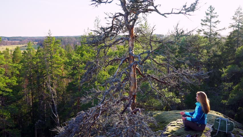 Young woman sitting in meditation pose on a mossy cliff surrounded by lush forest. Peaceful nature scene with sunlight filtering through trees. Mindfulness, solitude, mental well-being in outdoors.