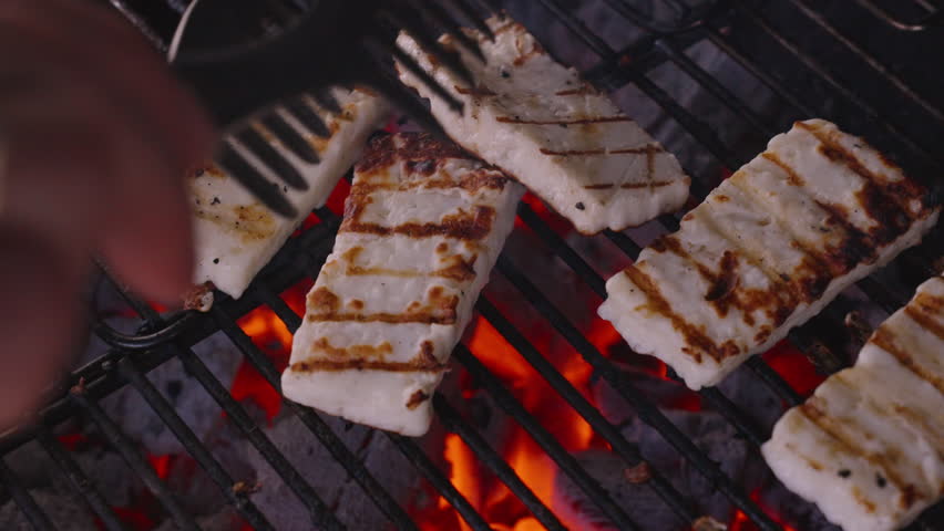 Halloumi Cheese Slices Being Carefully Flipped with Tongs on a Grill, Grill Marks Visible, One Slice Darkened, Over Glowing Charcoal, Angled Close-Up