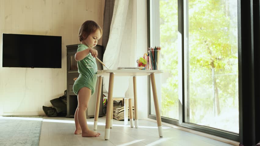 A child happily paints with a brush at a small table, opposite a large window in a small house in a bright room