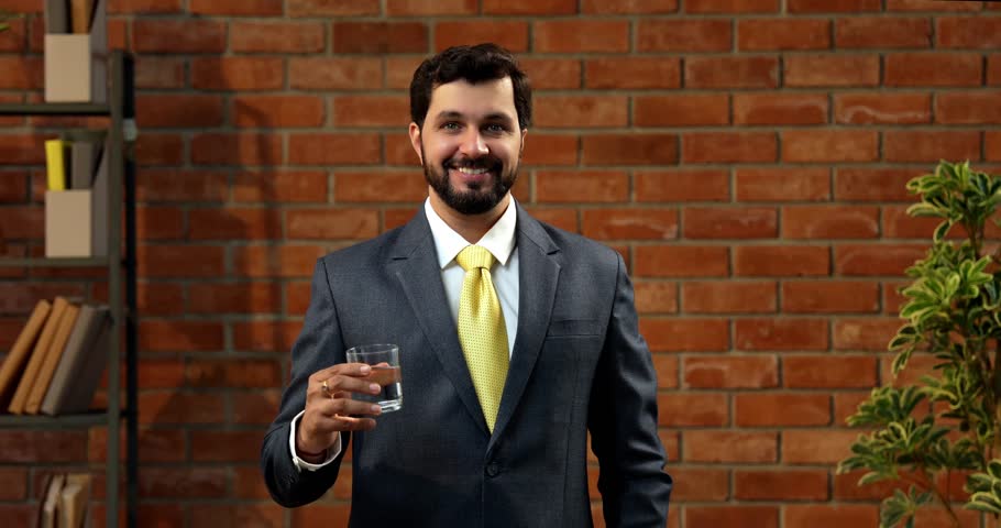 Drinking water from transparent glass, Indian businessman in formal wear highlights health and hydration. confident Asian CEO promotes timely water intake in cozy office with terracotta brick wall