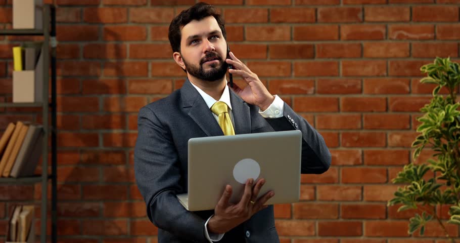 Indian businessman confidently speaking on smartphone while holding laptop, Asian CEO or entrepreneur in formal corporate attire with beard looks at computer screen in cozy office