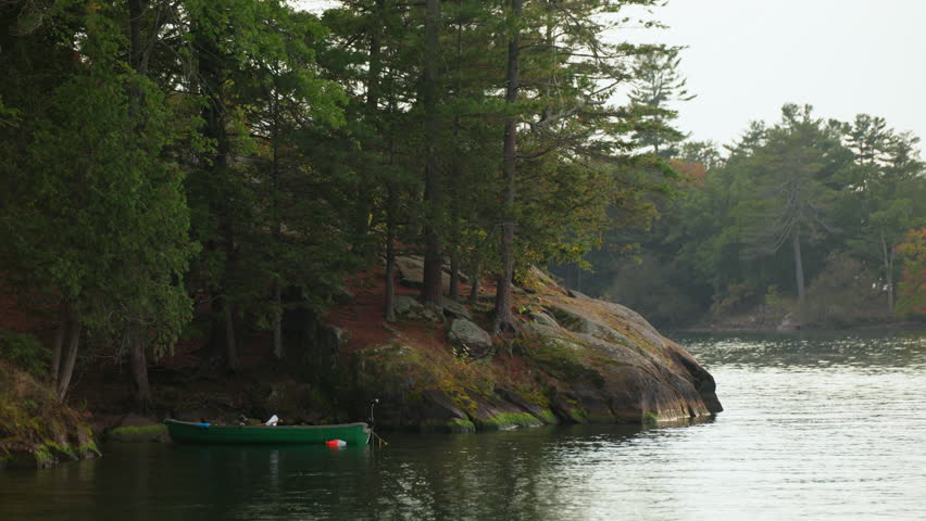 Handheld shot of a serene cove at Wellesley Island State Park, Fineview, NY, featuring a moored green rowboat by a rocky, forested riverbank.