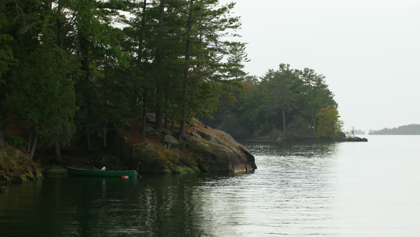 Handheld shot capturing a tranquil cove at Wellesley Island State Park, Fineview, NY, with a green rowboat moored by a forested, rocky shoreline.