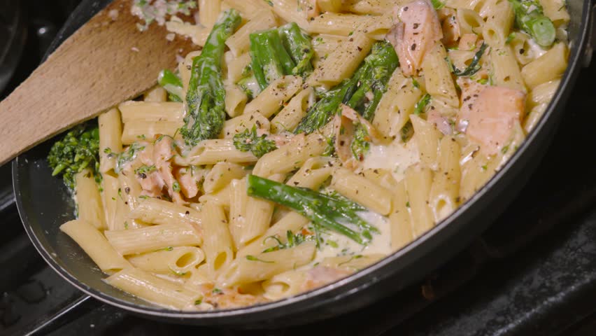 Top View Over Salmon and Broccoli Penne Italian Pasta Resting in Pan with Wooden Utensil with Cracked Black Pepper in Rich Creamy Sauce. Cooking Kitchen Seafood Healthy Eating Footage 4K.