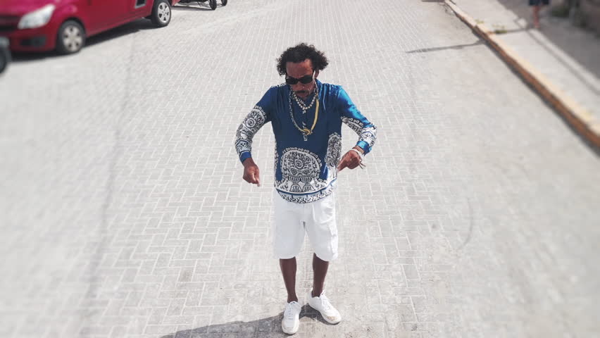 Afro Brazilian rap singer walks on the street, dances and sings. Drone camera flies away rapidly after a while and reveals the crowded beach and coastal town