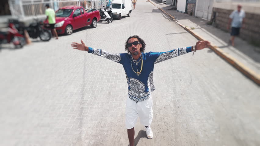 Afro Brazilian rap singer walks on the street, dances and sings. Drone camera flies away rapidly after a while and reveals the crowded beach and coastal town