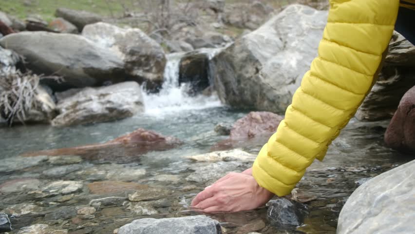 Female hiker touching pure mountain river stream, standing beside cascading waterfall, surrounded by rugged rocky landscape