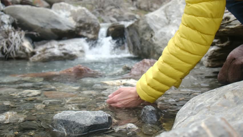 Female hiker touching pure mountain river stream, standing beside cascading waterfall, surrounded by rugged rocky landscape
