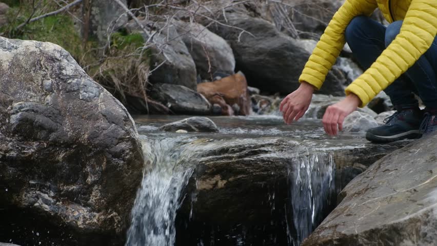 Female hiker touching pure mountain river stream, standing beside cascading waterfall, surrounded by rugged rocky landscape