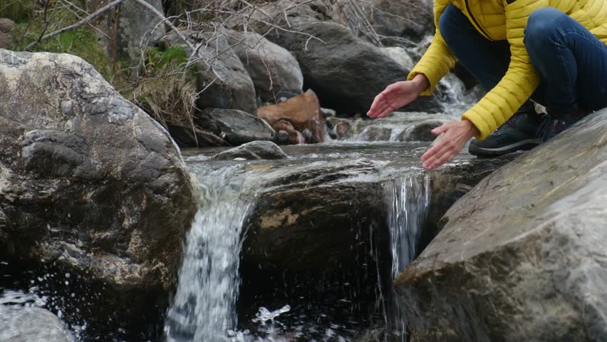 Female hiker touching pure mountain river stream, standing beside cascading waterfall, surrounded by rugged rocky landscape