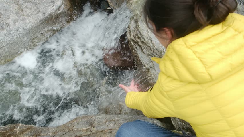 Female hiker touching pure mountain river stream, standing beside cascading waterfall, surrounded by rugged rocky landscape