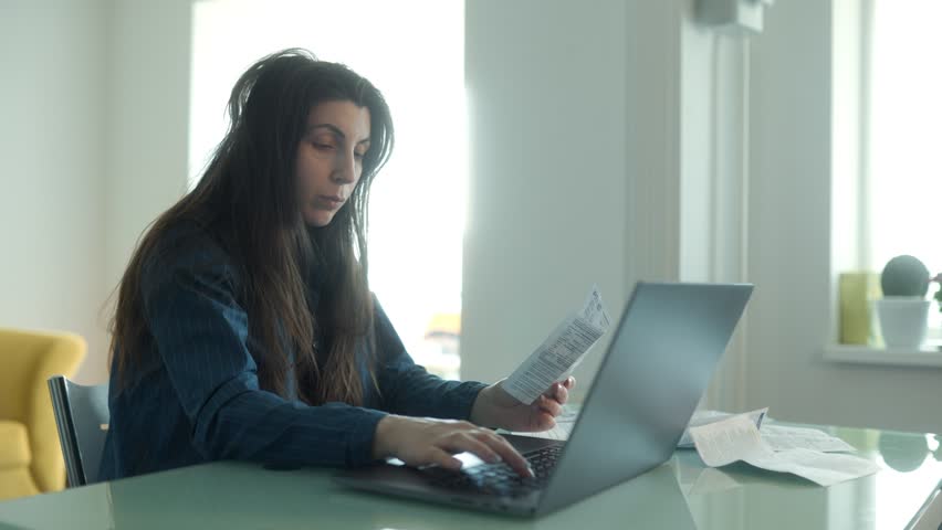 Focused woman managing online bill payments at home, surrounded by receipts and paperwork. She reviews financial documents while working on her laptop.