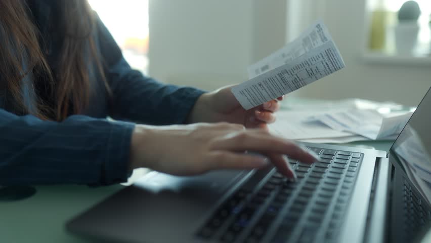 Close-up of a woman typing on a laptop while holding receipts, making online payments. The table is scattered with bills, reflecting a personal finance task. - Powered by Shutterstock - Get 15% off with code: PIKWIZARD15