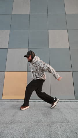 Vertical format video capturing expressive freestyle dancer in urban setting. Architectural tile wall, casual streetwear, strong pose, hip-hop influence, city atmosphere in full daylight. Freedom