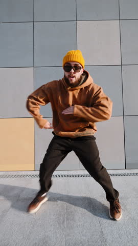 Vertical video confident young man dancing near geometric tile facade. Yellow sweatshirt, cargo pants, street-style performance, city lifestyle, youth culture captured in motion. Freedom urban