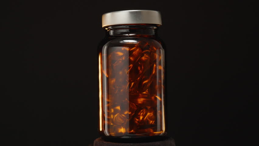 Looping 360-Degree Rotation of a Transparent Brown Glass Bottle Filled with Omega 3 Capsules Against a Dark Background with Warm Light Reflections