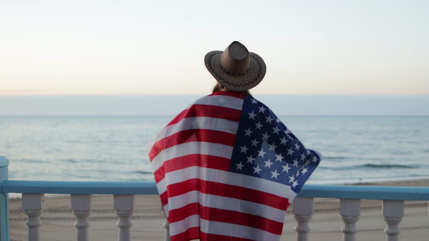 Woman standing proudly on sandy beach, waving united states flag with patriotic spirit during national celebration near ocean horizon.Usa celebrate 4th july. Independence day concept