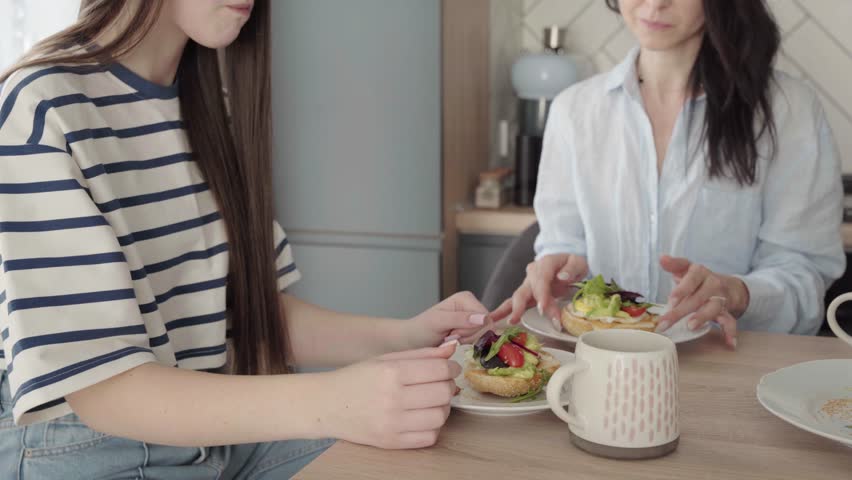 Teen girl and her mother in their 40s spending time together in modern kitchen, talking, laughing, drinking coffee, eating healthy sandwiches. Morning routine, natural light, lifestyle and bonding.