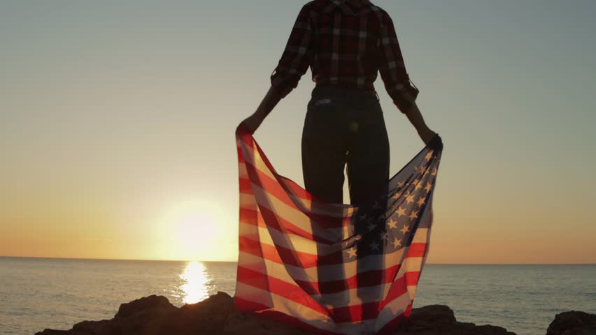 Female silhouette holding american flag, standing on rocky coastline during golden sunrise, embodying patriotic spirit with dramatic backlighting.Usa celebrate 4th july. Independence day concept - Powered by Shutterstock - Get 15% off with code: PIKWIZARD15