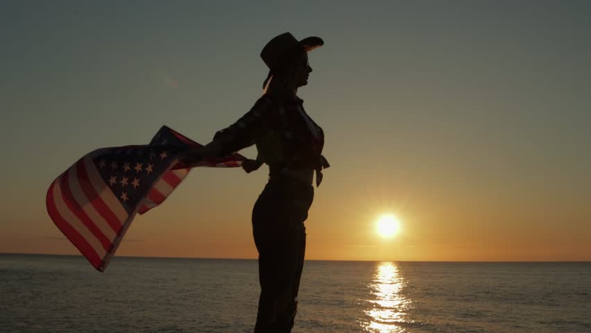 Silhouetted female wearing cowboy hat, waving american flag near ocean horizon during golden sunrise, embodying national pride and freedom.Usa celebrate 4th july. Independence day concept