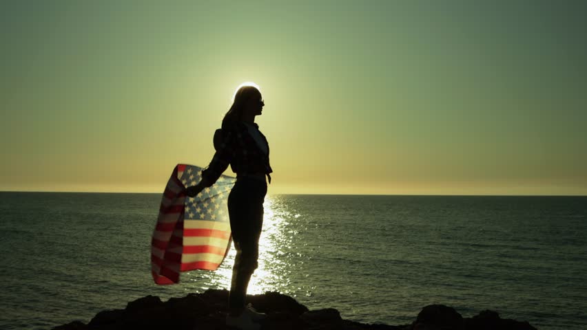 Patriotic female silhouette holding american flag, standing on rocky coastline during golden sunset, embodying national pride.Usa celebrate 4th july. Independence day concept